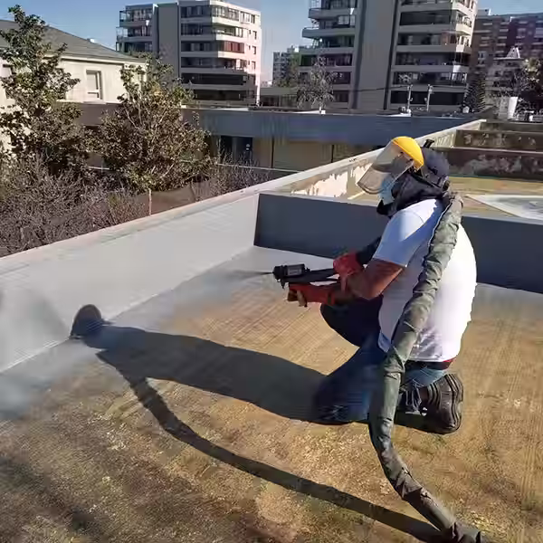 Construcción Operador con protector facial y EPP aplicando poliurea proyectada mediante pistola de alta presión sobre la losa expuesta de una azotea de hormigón en un edificio urbano.