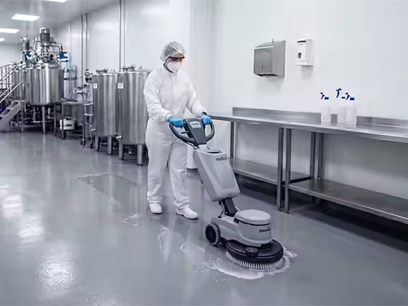 Una operaria con traje de protección blanco, mascarilla, gafas y cofia maneja una máquina fregadora industrial sobre un suelo gris brillante en una planta de producción limpia. Al fondo se observan tanques de acero inoxidable y mesas de trabajo relucientes.